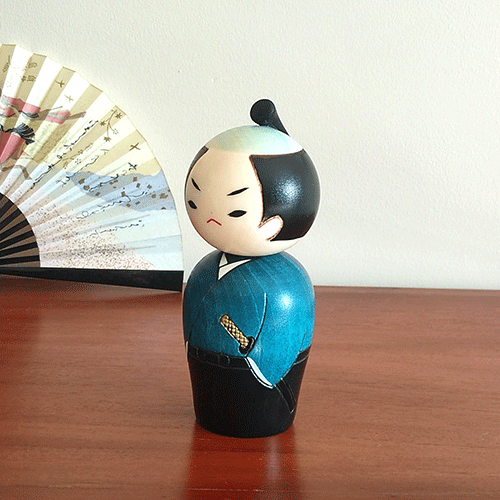 Handcrafted Samurai Kokeshi doll by Usaburo, dressed in a blue-green kimono and hakama, with a determined warrior’s expression and traditional chonmage topknot hairstyle, symbolizing samurai strength. Buy online at j-okini.com in Malta