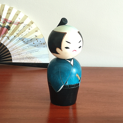 Handcrafted Samurai Kokeshi doll by Usaburo, dressed in a blue-green kimono and hakama, with a determined warrior’s expression and traditional chonmage topknot hairstyle, symbolizing samurai strength. Buy online at j-okini.com in Malta