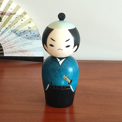 Handcrafted Samurai Kokeshi doll by Usaburo, dressed in a blue-green kimono and hakama, with a determined warrior’s expression and traditional chonmage topknot hairstyle, symbolizing samurai strength. Buy online at j-okini.com in Malta