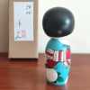 Hand-painted Kokeshi doll named Ryouhu, designed by artist Izumi, dressed in a blue kimono with morning glory flowers and a wind chime, symbolizing a cool Japanese summer breeze.