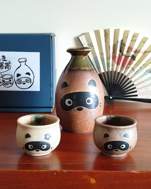 Sake set features an adorable Tanuki (raccoon dog) design, bringing a playful touch to your sake experience. The front of the sake bottle and cups showcase the face of the Tanuki, while the back reveals its tail, creating a delightful 360° design. The top portion is glazed in a rich green hue, while the unglazed bottom highlights the intricate craftsmanship of this set.. Made in Japan. Available at j-okini.com in Malta