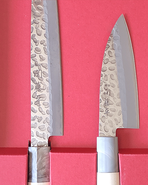 Gift masterpieces of Gifu and Seki of Japan. Yaxell is one of the major knife manufacturers in Japan since 1932, each Yaxell knife is a testament to the artistry and meticulous craftsmanship that has defined Japanese knife-making for generations, making them not just tools, but auspicious gifts that symbolize good fortune and prosperity. Made by Seki Tohei, and there are sign of 関藤平 on each knife. Blade It features a striking hammered design and provides comfortable cutting. Handle Sculpted from natural wood, designed to harmonize with your grip over time, becoming an extension of your culinary expression. This natural material not only adds to the knife's aesthetic but ensures durability and comfort with every use. This gift set includes: 1. 刺身包丁 Sashimi Bocho Overall length 330mm Blade length 205mm Weight: 79g Designed for precision, the Sashimi Bocho is the epitome of Japanese culinary artistry. Its long, slender blade is crafted to execute the most delicate and thin slices, perfect for sashimi, showcasing the knife's blend of functionality and elegance. 2.小出刃包丁 Small deba bocho Overall length 220mm Blade length 105mm Weight: 44g Specialized for handling smaller fish, this knife combines robustness and precision. Its compact design and sharp edge make it ideal for detailed work, from filleting to intricate cuts, embodying the spirit of traditional Japanese fish preparation.. Available at j-okini.com Malta