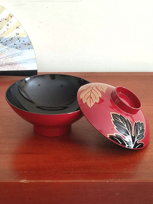 This elegant vintage Japanese lacquerware bowl, complete with a matching lid, is traditionally used for serving soup. The set of five was discovered carefully stored in a box, suggesting its origin from the Showa period (1926-1989). While the box is no longer available, the bowls themselves remain in pristine condition, showing no signs of wear and likely either unused or used only a few times. The bowl is adorned in a rich, traditional red lacquer, featuring a striking design of leaves. One leaf is black with delicate gold tracings, while the other gleams entirely in gold, adding a sophisticated touch to this timeless piece of Japanese craftsmanship. Available at j-okini.com in Malta