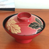 This elegant vintage Japanese lacquerware bowl, complete with a matching lid, is traditionally used for serving soup. The set of five was discovered carefully stored in a box, suggesting its origin from the Showa period (1926-1989). While the box is no longer available, the bowls themselves remain in pristine condition, showing no signs of wear and likely either unused or used only a few times. The bowl is adorned in a rich, traditional red lacquer, featuring a striking design of leaves. One leaf is black with delicate gold tracings, while the other gleams entirely in gold, adding a sophisticated touch to this timeless piece of Japanese craftsmanship. Available at j-okini.com in Malta