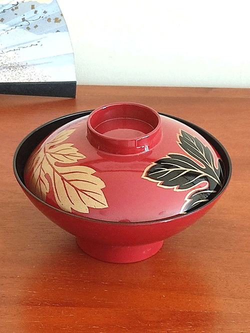 This elegant vintage Japanese lacquerware bowl, complete with a matching lid, is traditionally used for serving soup. The set of five was discovered carefully stored in a box, suggesting its origin from the Showa period (1926-1989). While the box is no longer available, the bowls themselves remain in pristine condition, showing no signs of wear and likely either unused or used only a few times. The bowl is adorned in a rich, traditional red lacquer, featuring a striking design of leaves. One leaf is black with delicate gold tracings, while the other gleams entirely in gold, adding a sophisticated touch to this timeless piece of Japanese craftsmanship. Available at j-okini.com in Malta