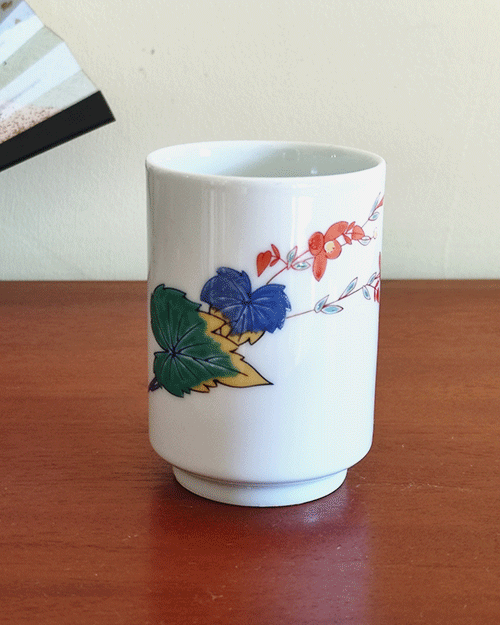 vintage teacup, crafted from white porcelain, features a  delicate hand-painted design of vibrant green, yellow, and blue leaves with a red flower gracefully extending toward the rim.. Available at j-okini.com in Malta