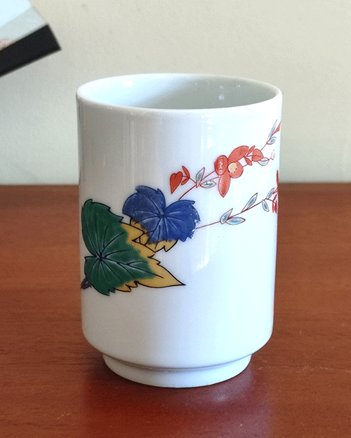 vintage teacup, crafted from white porcelain, features a  delicate hand-painted design of vibrant green, yellow, and blue leaves with a red flower gracefully extending toward the rim.. Available at j-okini.com in Malta