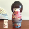 Modern Creative Kokeshi doll named Doshin, designed by artist Fujikawa Shoei, standing 20 cm tall and dressed in a pink and mauve kimono with red and white plum blossoms, symbolizing the innocence and purity of a child’s heart.. Buy it at j-okini.com in Malta