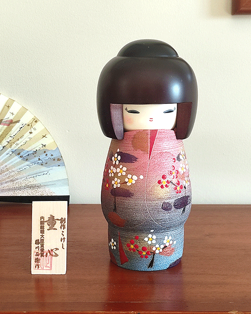 Modern Creative Kokeshi doll named Doshin, designed by artist Fujikawa Shoei, standing 20 cm tall and dressed in a pink and mauve kimono with red and white plum blossoms, symbolizing the innocence and purity of a child’s heart.. Buy it at j-okini.com in Malta