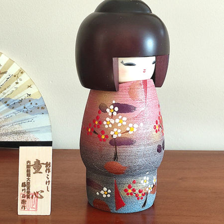 Modern Creative Kokeshi doll named Doshin, designed by artist Fujikawa Shoei, standing 20 cm tall and dressed in a pink and mauve kimono with red and white plum blossoms, symbolizing the innocence and purity of a child’s heart.. Buy it at j-okini.com in Malta