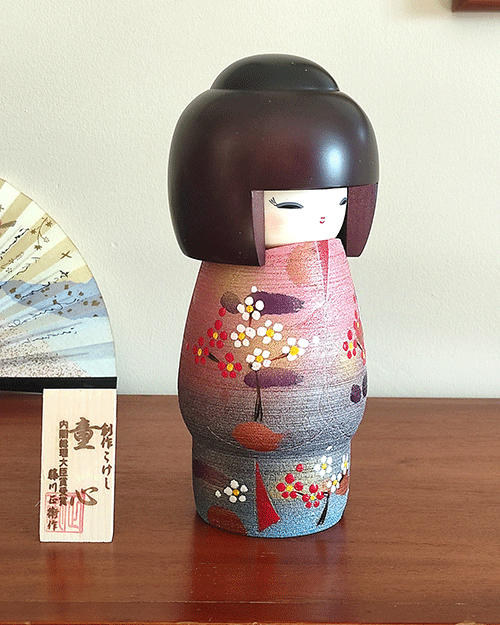 Modern Creative Kokeshi doll named Doshin, designed by artist Fujikawa Shoei, standing 20 cm tall and dressed in a pink and mauve kimono with red and white plum blossoms, symbolizing the innocence and purity of a child’s heart.. Buy it at j-okini.com in Malta