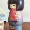 Modern Creative Kokeshi doll named Doshin, designed by artist Fujikawa Shoei, standing 20 cm tall and dressed in a pink and mauve kimono with red and white plum blossoms, symbolizing the innocence and purity of a child’s heart.. Buy it at j-okini.com in Malta