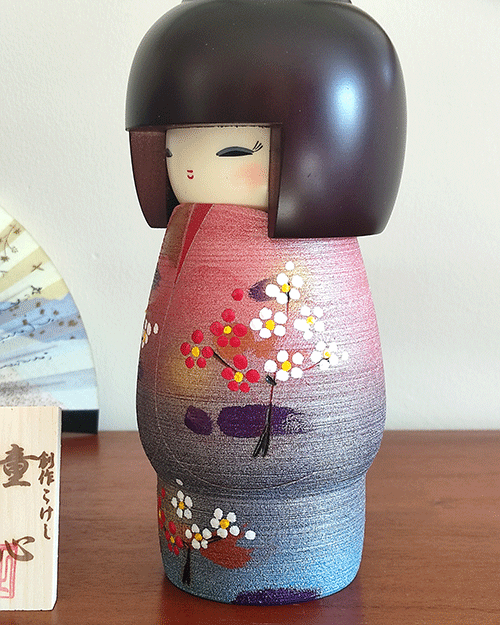 Modern Creative Kokeshi doll named Doshin, designed by artist Fujikawa Shoei, standing 20 cm tall and dressed in a pink and mauve kimono with red and white plum blossoms, symbolizing the innocence and purity of a child’s heart.. Buy it at j-okini.com in Malta