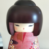 Modern Creative Kokeshi doll named Doshin, designed by artist Fujikawa Shoei, standing 20 cm tall and dressed in a pink and mauve kimono with red and white plum blossoms, symbolizing the innocence and purity of a child’s heart.. Buy it at j-okini.com in Malta