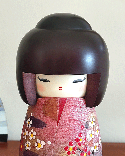 Modern Creative Kokeshi doll named Doshin, designed by artist Fujikawa Shoei, standing 20 cm tall and dressed in a pink and mauve kimono with red and white plum blossoms, symbolizing the innocence and purity of a child’s heart.. Buy it at j-okini.com in Malta