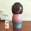 Modern Creative Kokeshi doll named Doshin, designed by artist Fujikawa Shoei, standing 20 cm tall and dressed in a pink and mauve kimono with red and white plum blossoms, symbolizing the innocence and purity of a child’s heart.. Buy it at j-okini.com in Malta