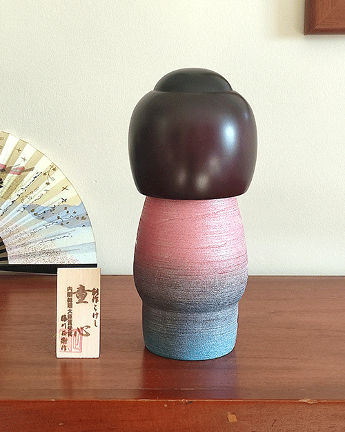 Modern Creative Kokeshi doll named Doshin, designed by artist Fujikawa Shoei, standing 20 cm tall and dressed in a pink and mauve kimono with red and white plum blossoms, symbolizing the innocence and purity of a child’s heart.. Buy it at j-okini.com in Malta