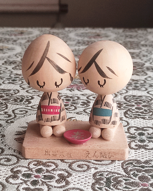 A pair of vintage wooden dolls from Izumo Taisha Shrine in Seiza position, representing a boy and girl. Hand-painted with traditional kimonos and seated with a red sake cup inscribed with "寿" (kotobuki). A faint craftsman's stamp is next to the girl on the wooden base. Available at j-okini.com in Malta