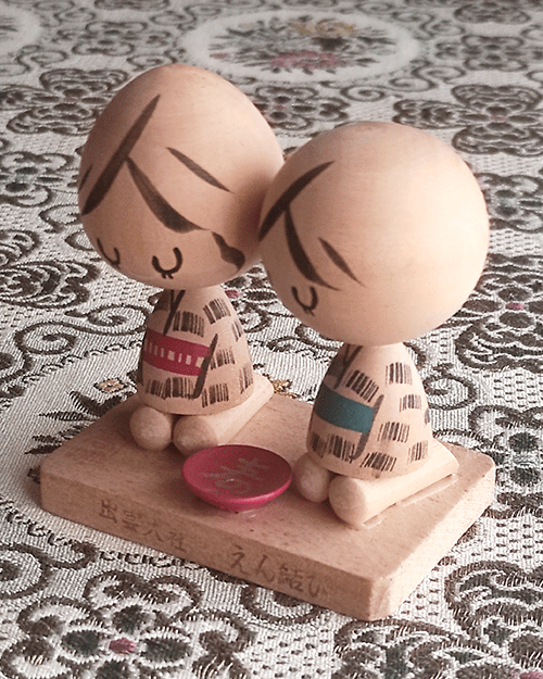 A pair of vintage wooden dolls from Izumo Taisha Shrine in Seiza position, representing a boy and girl. Hand-painted with traditional kimonos and seated with a red sake cup inscribed with "寿" (kotobuki). A faint craftsman's stamp is next to the girl on the wooden base.