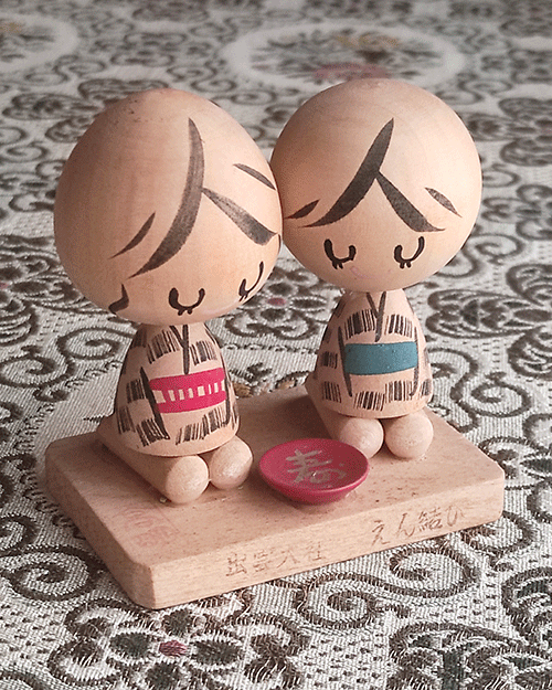 A pair of vintage wooden dolls from Izumo Taisha Shrine in Seiza position, representing a boy and girl. Hand-painted with traditional kimonos and seated with a red sake cup inscribed with "寿" (kotobuki). A faint craftsman's stamp is next to the girl on the wooden base.