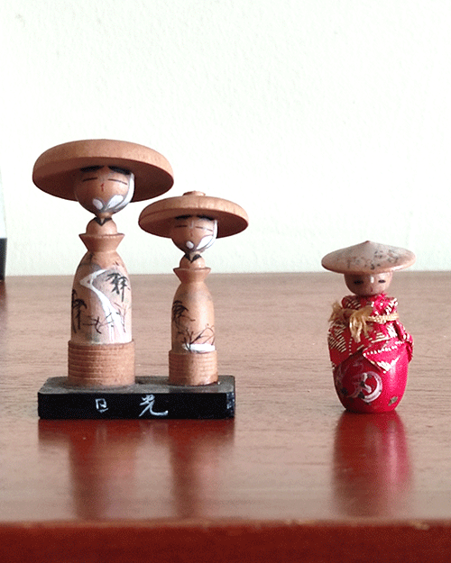 Two small vintage Japanese folk dolls from the Showa era: one in an orange kimono with a hat, and a wooden father-and-son pair standing on a wooden board with hand-painted details and traditional hats from Nikko, Tochigi Prefecture. Available at j-okini.com in Malta