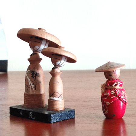 Two small vintage Japanese folk dolls from the Showa era: one in an orange kimono with a hat, and a wooden father-and-son pair standing on a wooden board with hand-painted details and traditional hats from Nikko, Tochigi Prefecture. Available at j-okini.com in Malta