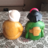 Vintage Hyottoko and Okame pottery ornaments from the Showa era, Hyottoko in a mustard kimono and Okame in a green kimono with red apron, approx. 15 cm tall, symbols of fire protection and good fortune. Available at j-okini.com in Malta