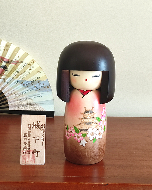 Kokeshi doll named Jokamachi featuring a beautiful depiction of a castle and delicate cherry blossoms on a soft pink kimono, symbolizing the elegance and history of Japanese heritage.. Buy it online at j-okini.com in Malta