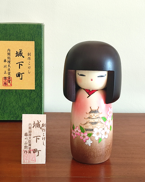 Kokeshi doll named Jokamachi featuring a beautiful depiction of a castle and delicate cherry blossoms on a soft pink kimono, symbolizing the elegance and history of Japanese heritage.. Buy it online at j-okini.com in Malta