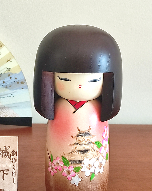 Kokeshi doll named Jokamachi featuring a beautiful depiction of a castle and delicate cherry blossoms on a soft pink kimono, symbolizing the elegance and history of Japanese heritage.. Buy it online at j-okini.com in Malta