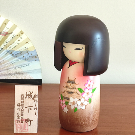 Kokeshi doll named Jokamachi featuring a beautiful depiction of a castle and delicate cherry blossoms on a soft pink kimono, symbolizing the elegance and history of Japanese heritage.. Buy it online at j-okini.com in Malta