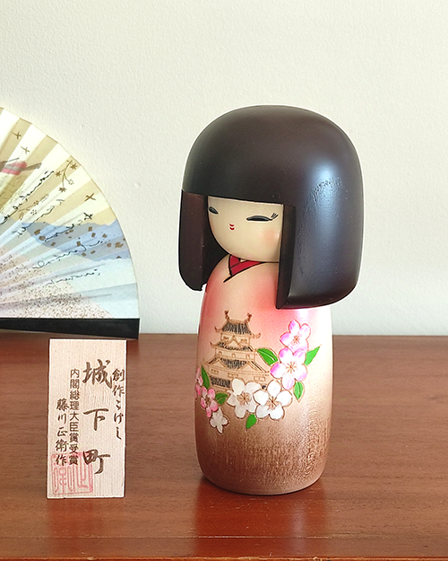 Kokeshi doll named Jokamachi featuring a beautiful depiction of a castle and delicate cherry blossoms on a soft pink kimono, symbolizing the elegance and history of Japanese heritage.. Buy it online at j-okini.com in Malta