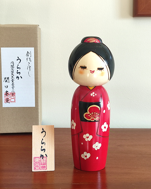 Modern Creative Kokeshi doll named Uraraka, crafted by award-winning artist Sekiguchi Toua, wearing a vibrant red kimono with plum blossom patterns, a black obi with matching design, and a plum blossom Kanzashi hair ornament in a stylish bun.. Buy it online at j-okini.com in Malta