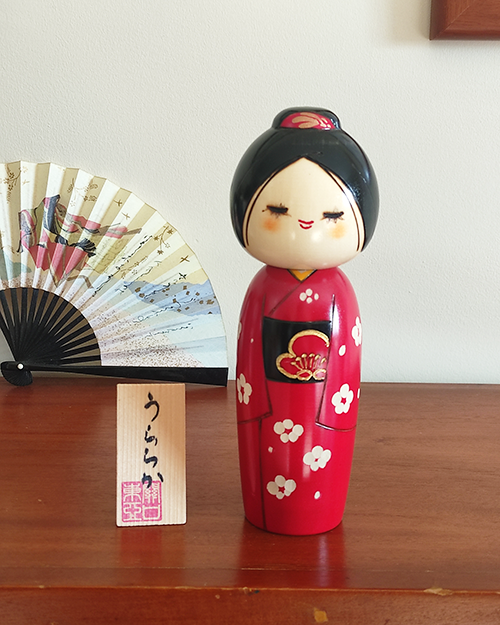 Modern Creative Kokeshi doll named Uraraka, crafted by award-winning artist Sekiguchi Toua, wearing a vibrant red kimono with plum blossom patterns, a black obi with matching design, and a plum blossom Kanzashi hair ornament in a stylish bun.. Buy it online at j-okini.com in Malta