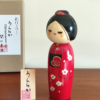 Modern Creative Kokeshi doll named Uraraka, crafted by award-winning artist Sekiguchi Toua, wearing a vibrant red kimono with plum blossom patterns, a black obi with matching design, and a plum blossom Kanzashi hair ornament in a stylish bun.. Buy it online at j-okini.com in Malta