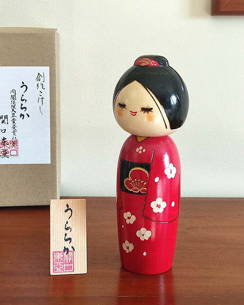 Modern Creative Kokeshi doll named Uraraka, crafted by award-winning artist Sekiguchi Toua, wearing a vibrant red kimono with plum blossom patterns, a black obi with matching design, and a plum blossom Kanzashi hair ornament in a stylish bun.. Buy it online at j-okini.com in Malta