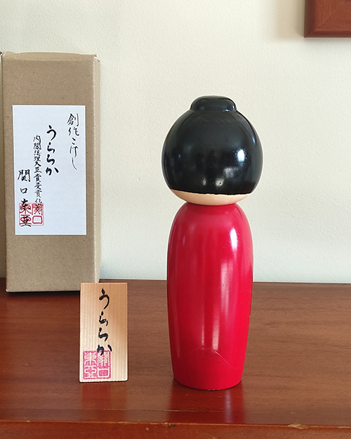 Modern Creative Kokeshi doll named Uraraka, crafted by award-winning artist Sekiguchi Toua, wearing a vibrant red kimono with plum blossom patterns, a black obi with matching design, and a plum blossom Kanzashi hair ornament in a stylish bun.. Buy it online at j-okini.com in Malta