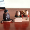 Two vintage wooden dolls from the Showa era: one shaped like an aubergine with a sweet face resting on a wooden leaf, and another pair of donguri dolls seated on wooden geta sandals, featuring hand-painted details and a mushroom accent.