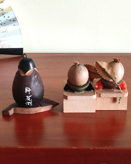 Two vintage wooden dolls from the Showa era: one shaped like an aubergine with a sweet face resting on a wooden leaf, and another pair of donguri dolls seated on wooden geta sandals, featuring hand-painted details and a mushroom accent.