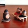 Two vintage wooden dolls from the Showa era: one shaped like an aubergine with a sweet face resting on a wooden leaf, and another pair of donguri dolls seated on wooden geta sandals, featuring hand-painted details and a mushroom accent.