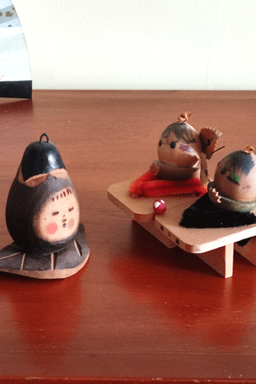 Two vintage wooden dolls from the Showa era: one shaped like an aubergine with a sweet face resting on a wooden leaf, and another pair of donguri dolls seated on wooden geta sandals, featuring hand-painted details and a mushroom accent.