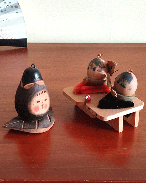 Two vintage wooden dolls from the Showa era: one shaped like an aubergine with a sweet face resting on a wooden leaf, and another pair of donguri dolls seated on wooden geta sandals, featuring hand-painted details and a mushroom accent.