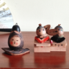 Two vintage wooden dolls from the Showa era: one shaped like an aubergine with a sweet face resting on a wooden leaf, and another pair of donguri dolls seated on wooden geta sandals, featuring hand-painted details and a mushroom accent. Available at j-okini.com in Malta