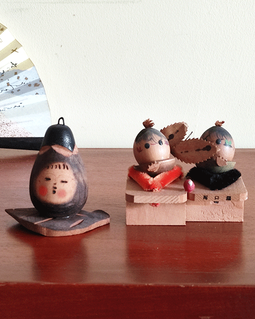 Two vintage wooden dolls from the Showa era: one shaped like an aubergine with a sweet face resting on a wooden leaf, and another pair of donguri dolls seated on wooden geta sandals, featuring hand-painted details and a mushroom accent. Available at j-okini.com in Malta