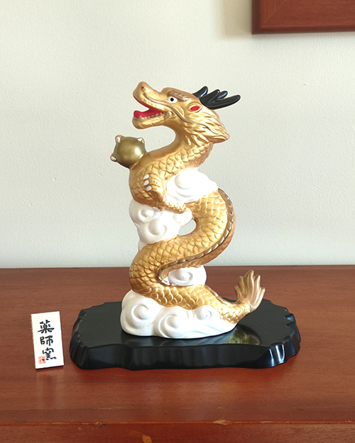 A golden dragon figurine standing 19cm tall, depicted soaring into the sky amidst white clouds, holding a "Hoju" (sacred jewel) in its claw. The dragon's dynamic pose symbolizes prosperity and improved luck, while the black lacquered base adds a touch of elegance. Gift idea from j-okini.com in Malta