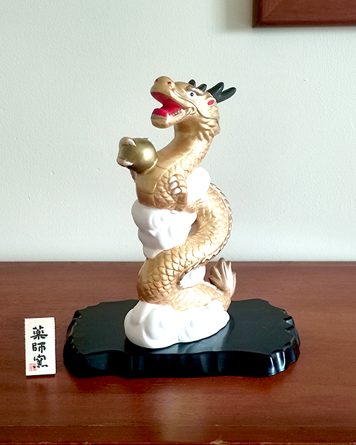 A golden dragon figurine standing 19cm tall, depicted soaring into the sky amidst white clouds, holding a "Hoju" (sacred jewel) in its claw. The dragon's dynamic pose symbolizes prosperity and improved luck, while the black lacquered base adds a touch of elegance. Gift idea from j-okini.com in Malta