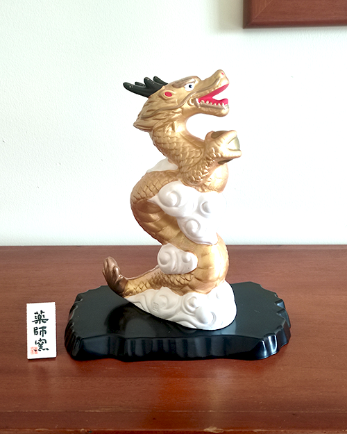 A golden dragon figurine standing 19cm tall, depicted soaring into the sky amidst white clouds, holding a "Hoju" (sacred jewel) in its claw. The dragon's dynamic pose symbolizes prosperity and improved luck, while the black lacquered base adds a touch of elegance. Gift idea from j-okini.com in Malta