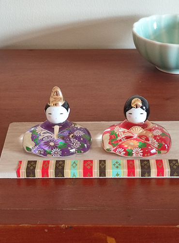 Porcelain and glass Hina dolls for Japan’s Girl’s Festival, with the Emperor in purple and the Empress in red, each decorated with delicate floral designs on a wooden platform.. Buy it in Malta at j-okini.com