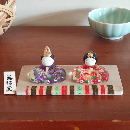 Porcelain and glass Hina dolls for Japan’s Girl’s Festival, with the Emperor in purple and the Empress in red, each decorated with delicate floral designs on a wooden platform.. Buy it in Malta at j-okini.com