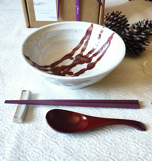 A beautifully curated Japanese ramen dining set featuring a handcrafted white ramen bowl with red accents, rose red wooden spoon, hexagonal chopsticks, and a ceramic chopstick rest, elegantly displayed on a white textured background. j-okini.com in Malta